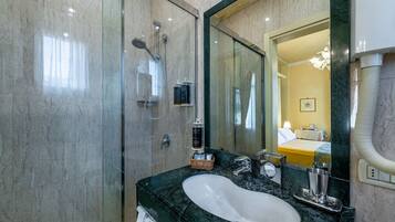 A modern bathroom with a glass shower enclosure, a marble sink, and a large mirror reflecting a bedroom with a bed and a nightstand.