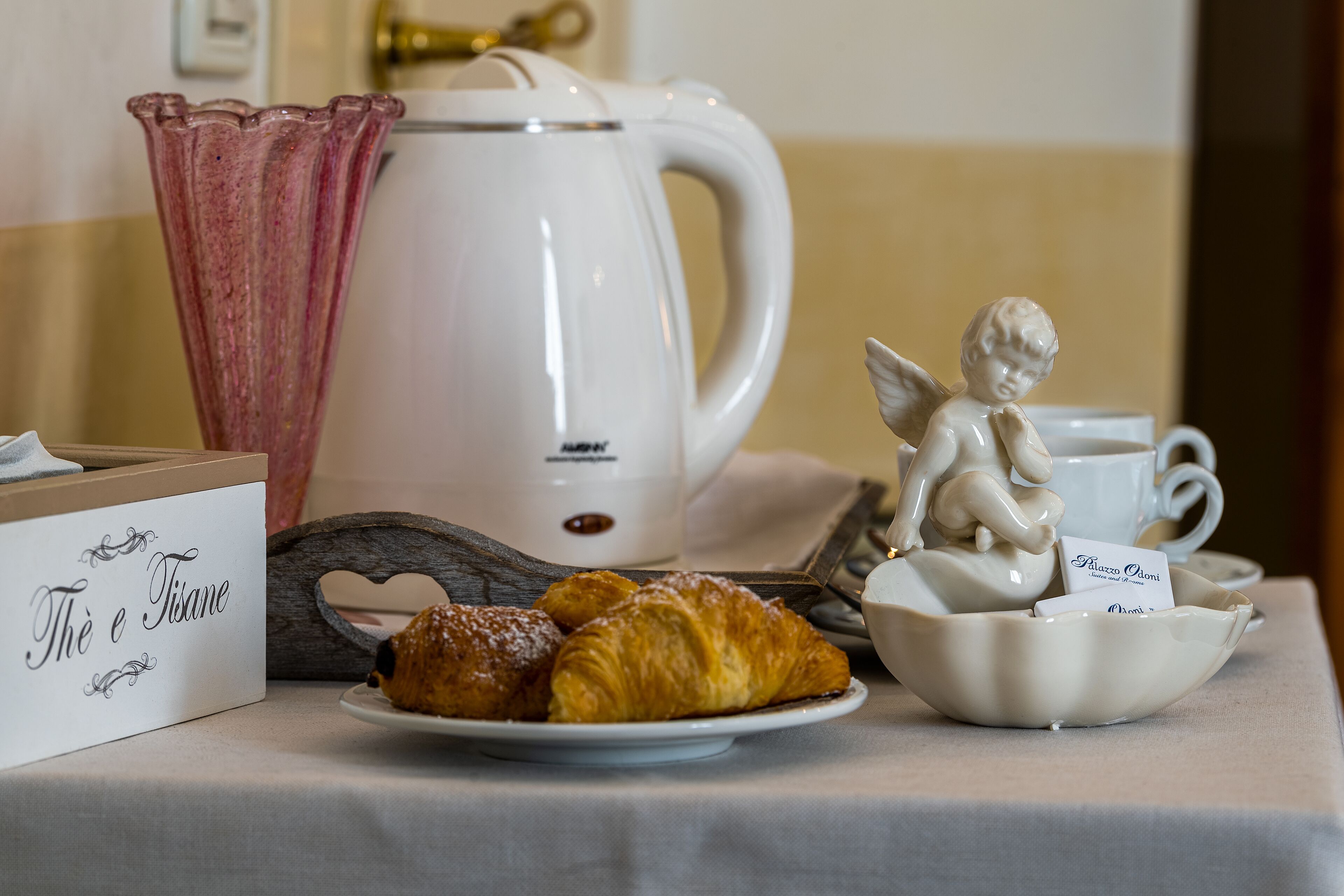 A breakfast setup with croissants, a coffee maker, a teapot, and a decorative angel figurine.