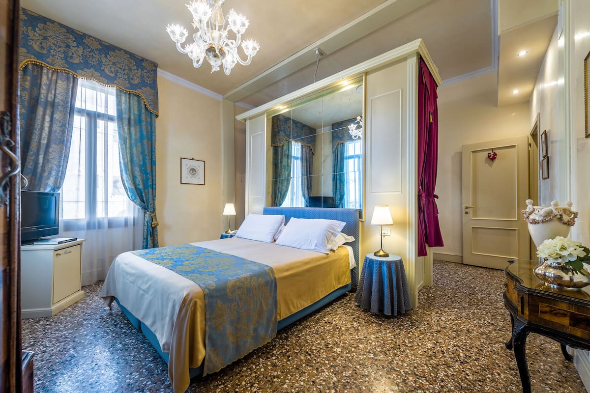 A hotel room with a large bed, a television, a mirror, and a chandelier.