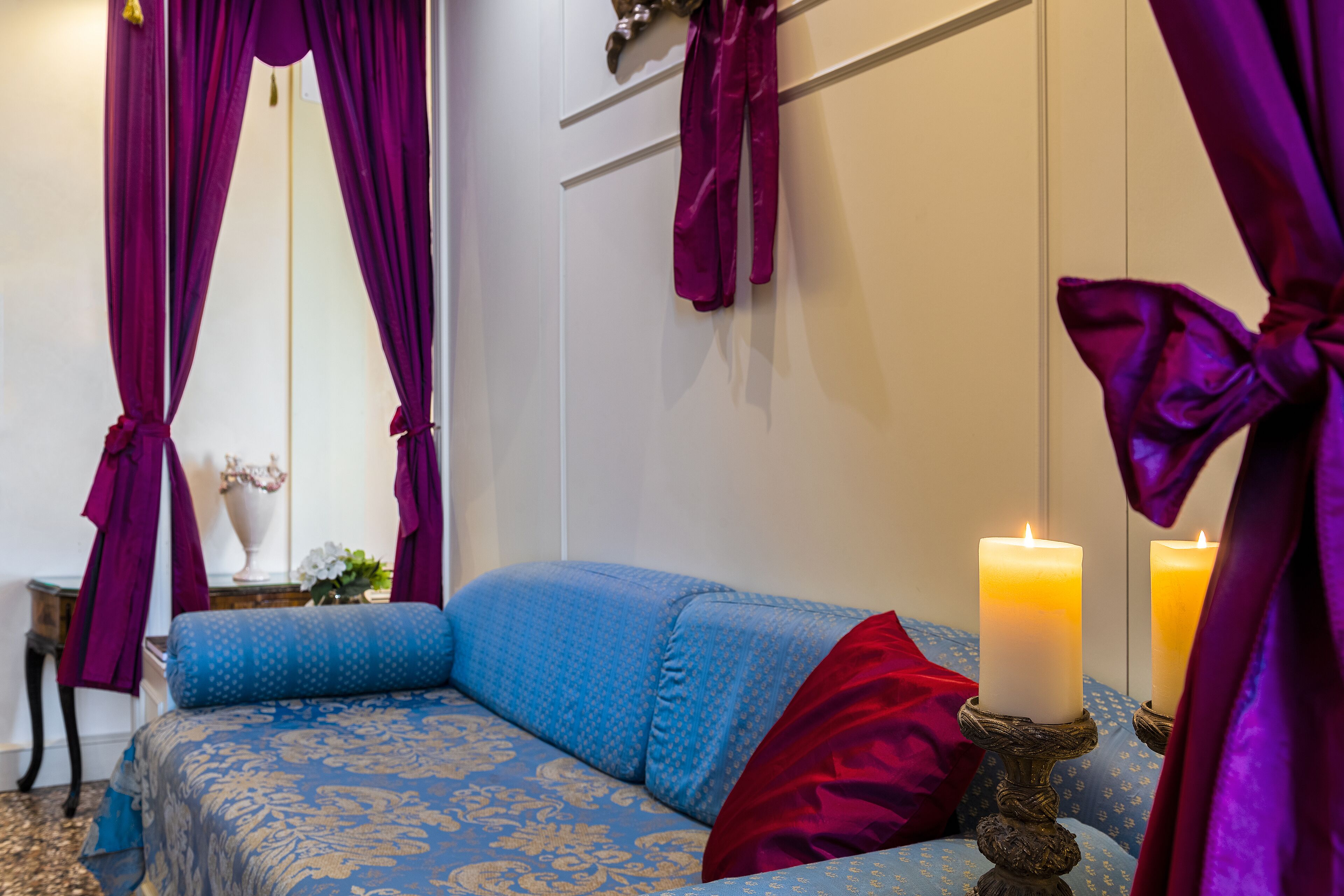 A bedroom with a blue patterned bedspread, two candles on a decorative stand, and purple curtains.