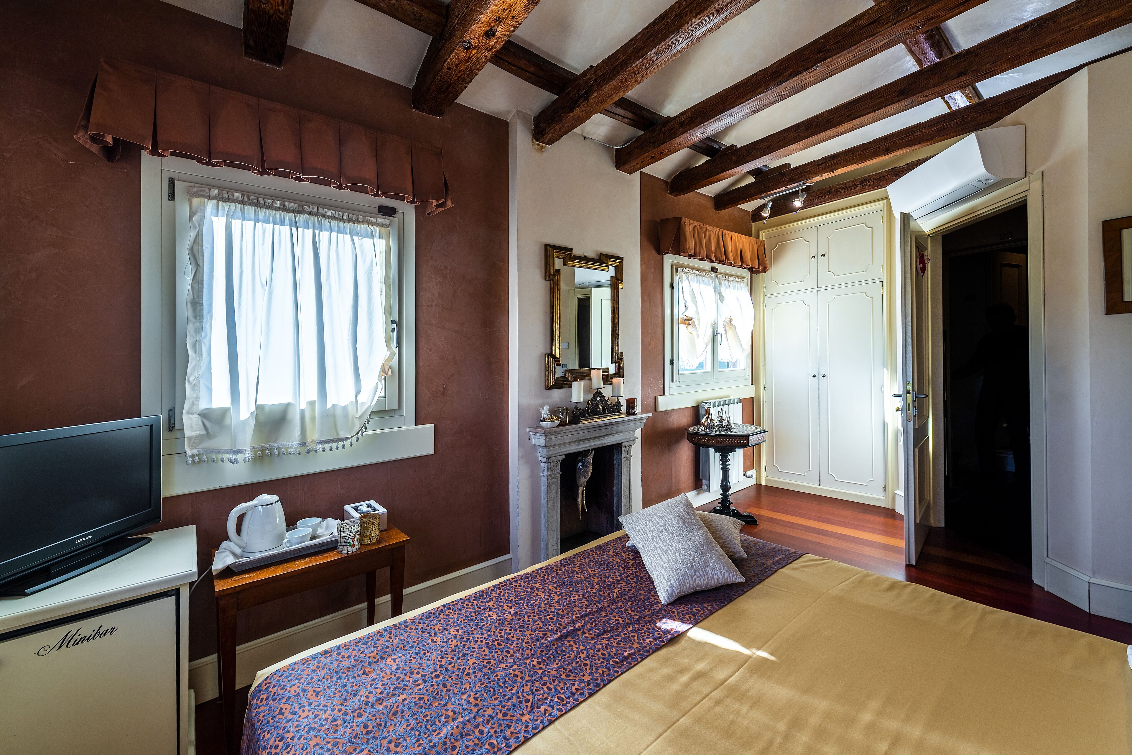 A hotel room with a bed, a television, a desk with a kettle, a fireplace, and a mirror.