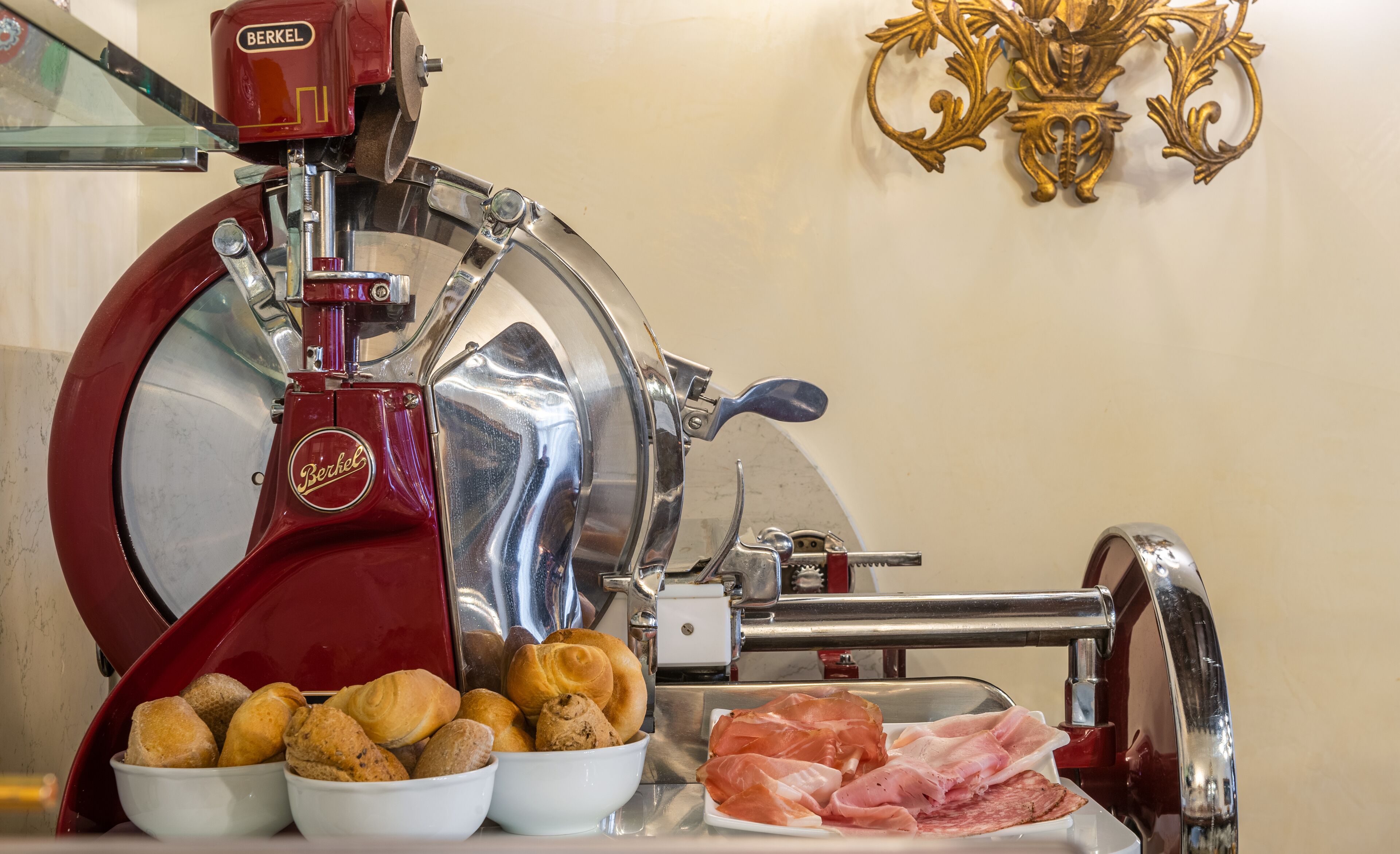 A meat slicer with a red base and a shiny metallic top, surrounded by bowls of bread and sliced meat.