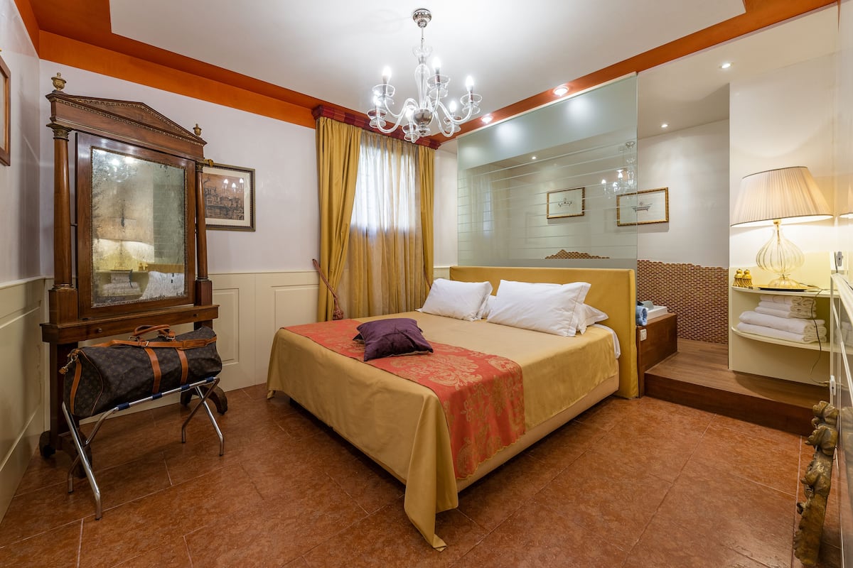 A hotel room with a bed, a bedside table, a mirror, a chandelier, and a suitcase on a trolley.