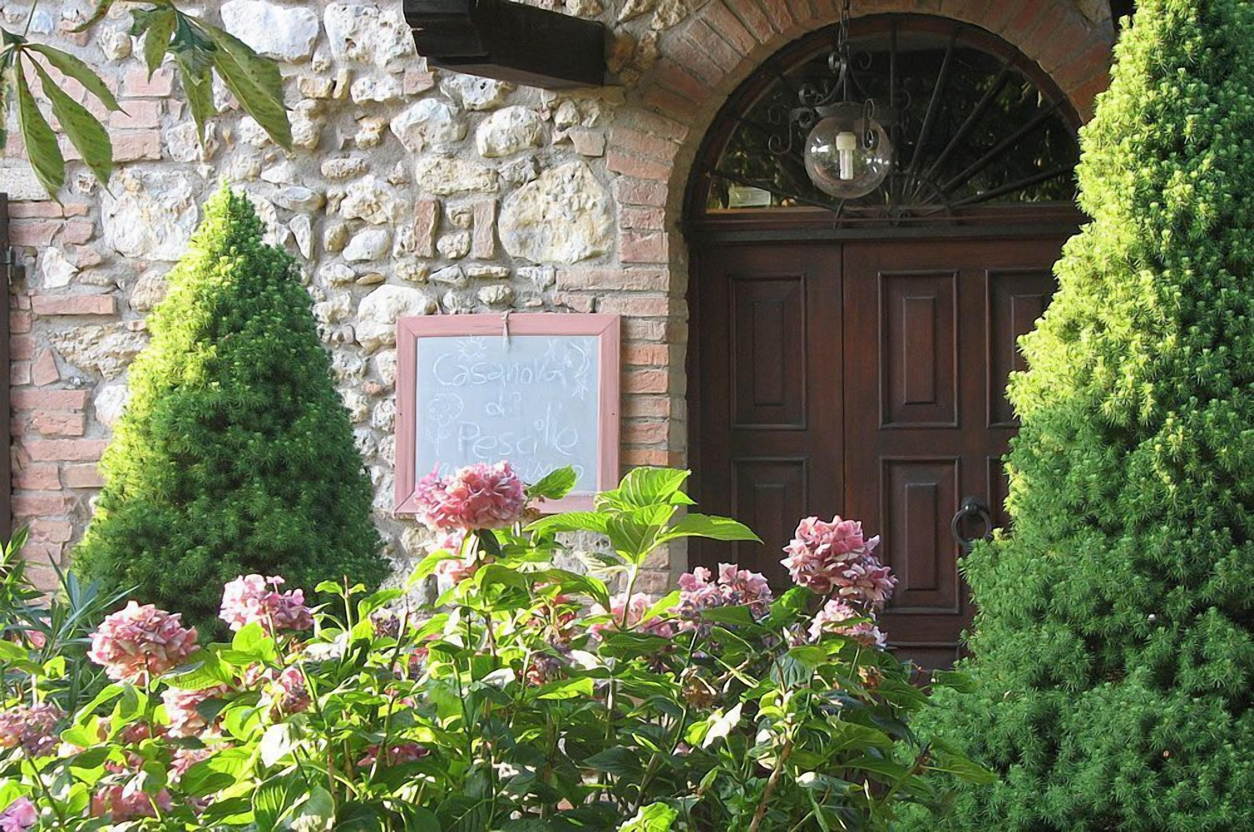 Un edificio in pietra con una porta di legno, due alberi conici e fiori rosa in primo piano.