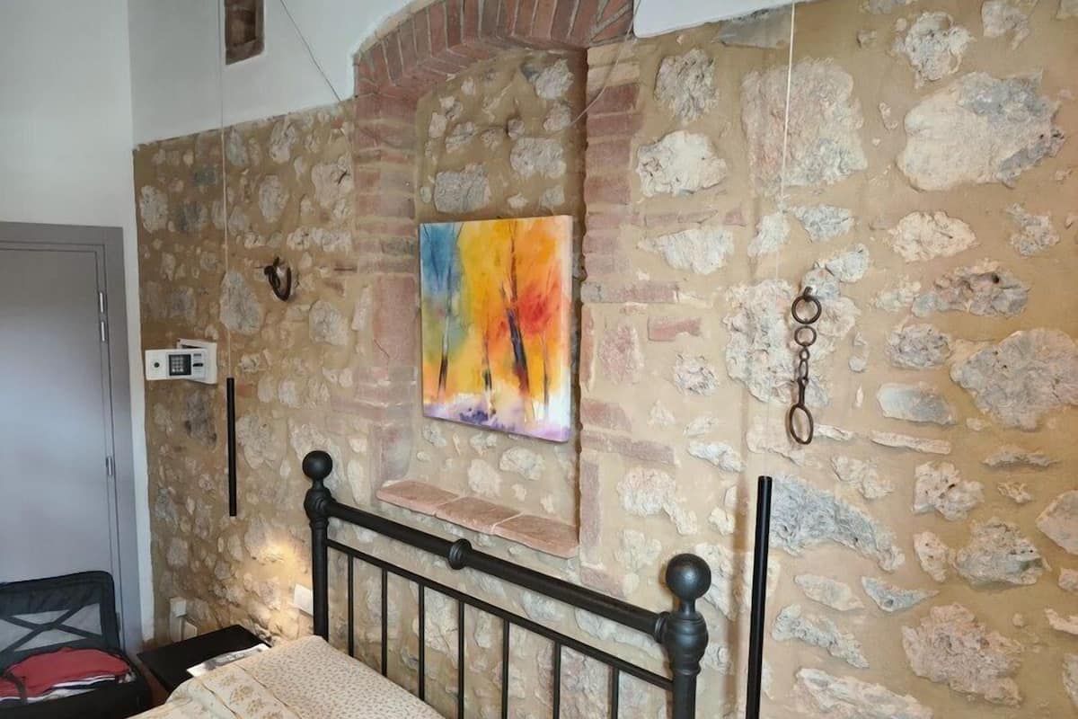 A bedroom with a stone wall, a bed with a black metal frame, a colorful abstract painting, and a small window.