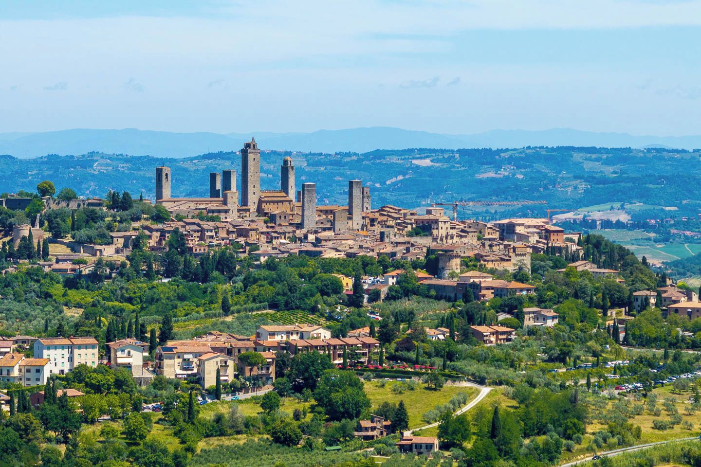 A historic town with medieval architecture, including towers and a fortified wall, surrounded by lush greenery and rolling hills.