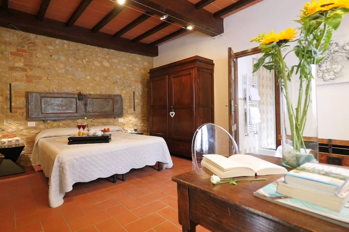 A bedroom with a bed, a wooden wardrobe, a bedside table with books, a vase with flowers, and a stone wall.