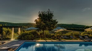 Un área de piscina con sillones de descanso y sombrillas, rodeada de exuberante vegetación y colinas.