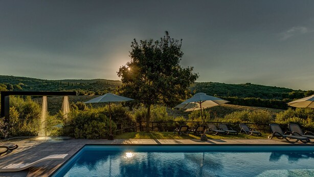 A pool area with lounge chairs and umbrellas, surrounded by lush greenery and hills. - Casanova di Pescille (San Gimignano)