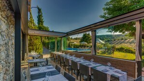 Un área de comedor al aire libre con mesas dispuestas para dos, con vista a una piscina y jardín.