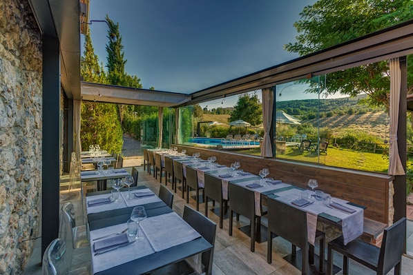 Un área de comedor al aire libre con mesas dispuestas para dos, con vista a una piscina y jardín.