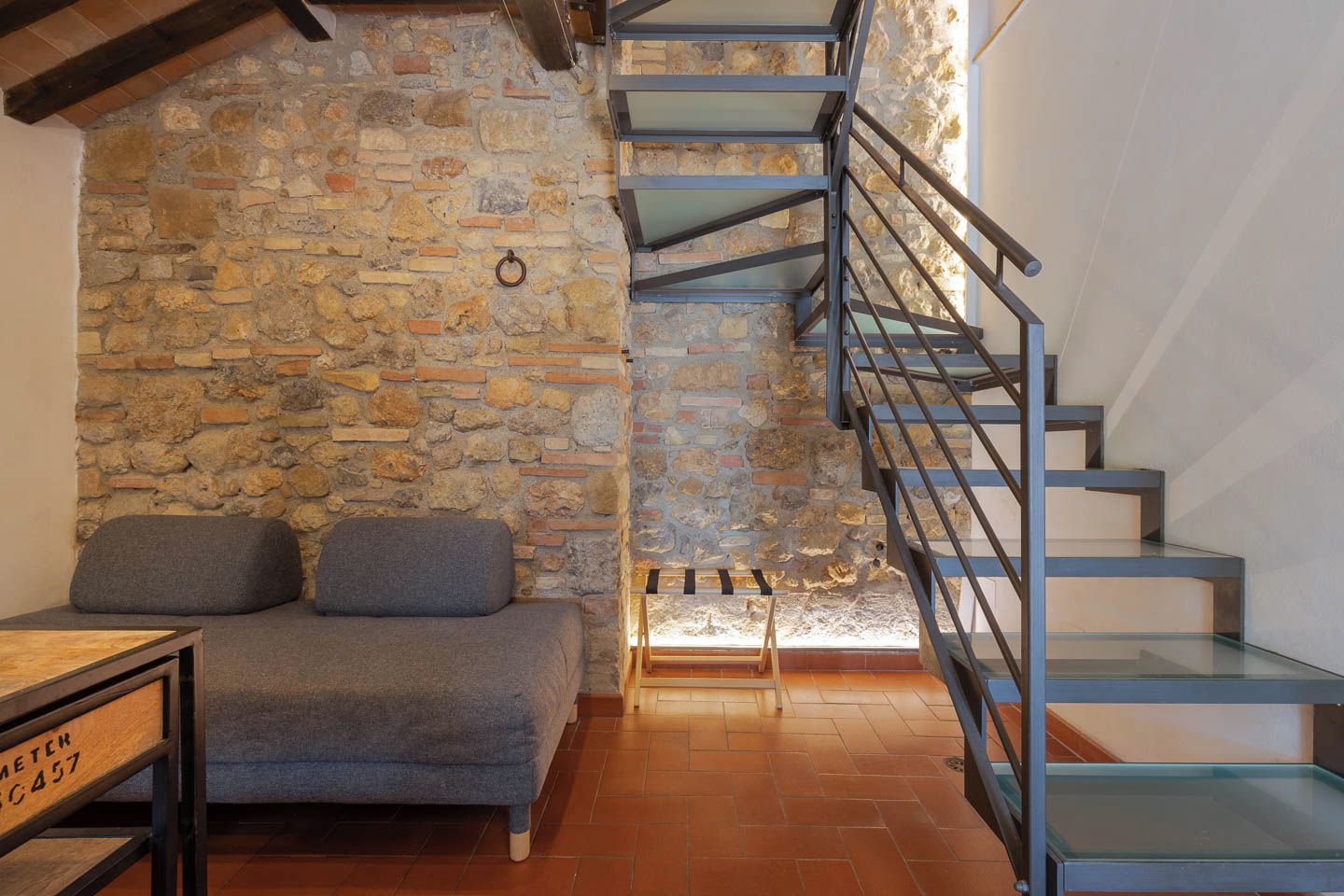 A room with a stone wall, a grey sofa, a wooden table, and a glass staircase.