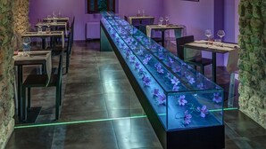 Un restaurante con una larga mesa mostrando flores moradas, cuadros de paisajes enmarcados en las paredes y muros de piedra.