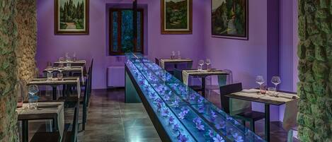 A restaurant with a long table displaying purple flowers, framed landscape paintings on the walls, and stone walls.