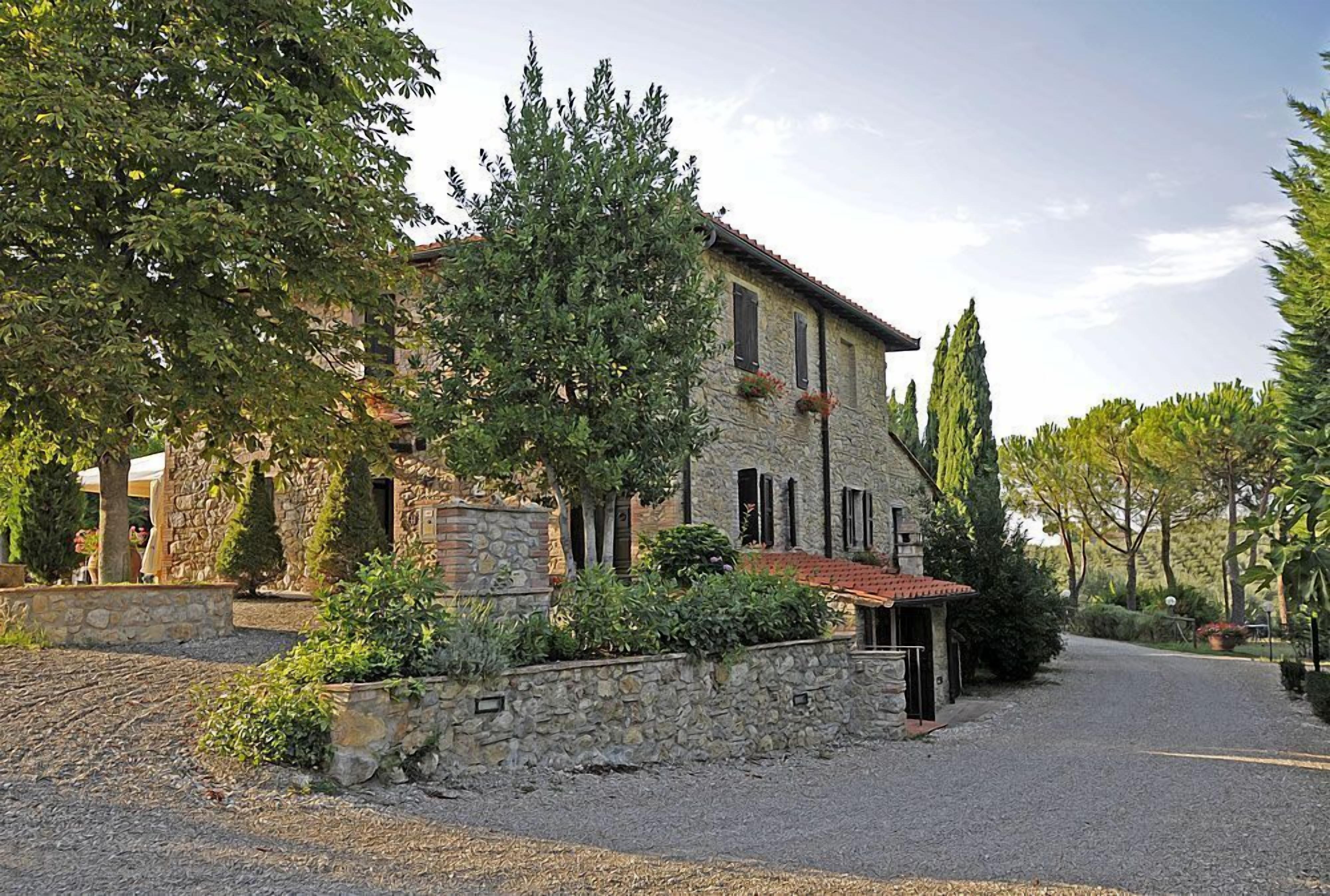A stone building with a red-tiled roof, surrounded by greenery and a gravel driveway.