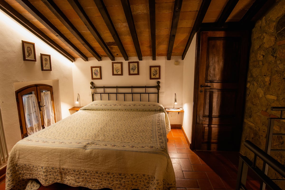 A bedroom with a wooden ceiling, a bed with a floral bedspread, a nightstand with a lamp, and a stone wall.
