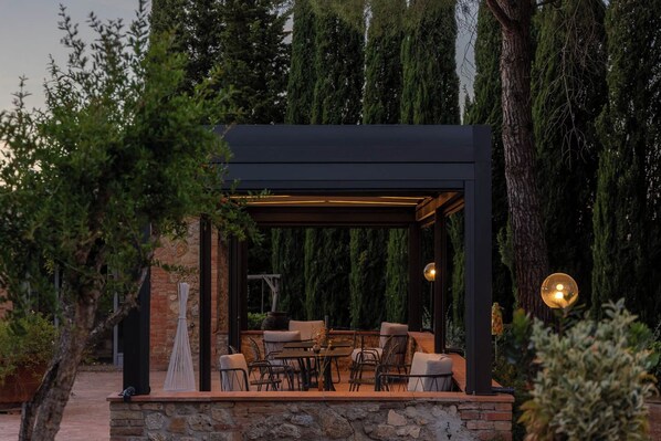 An outdoor seating area with a pergola, stone walls, and a view of tall trees. - Casanova di Pescille (San Gimignano)