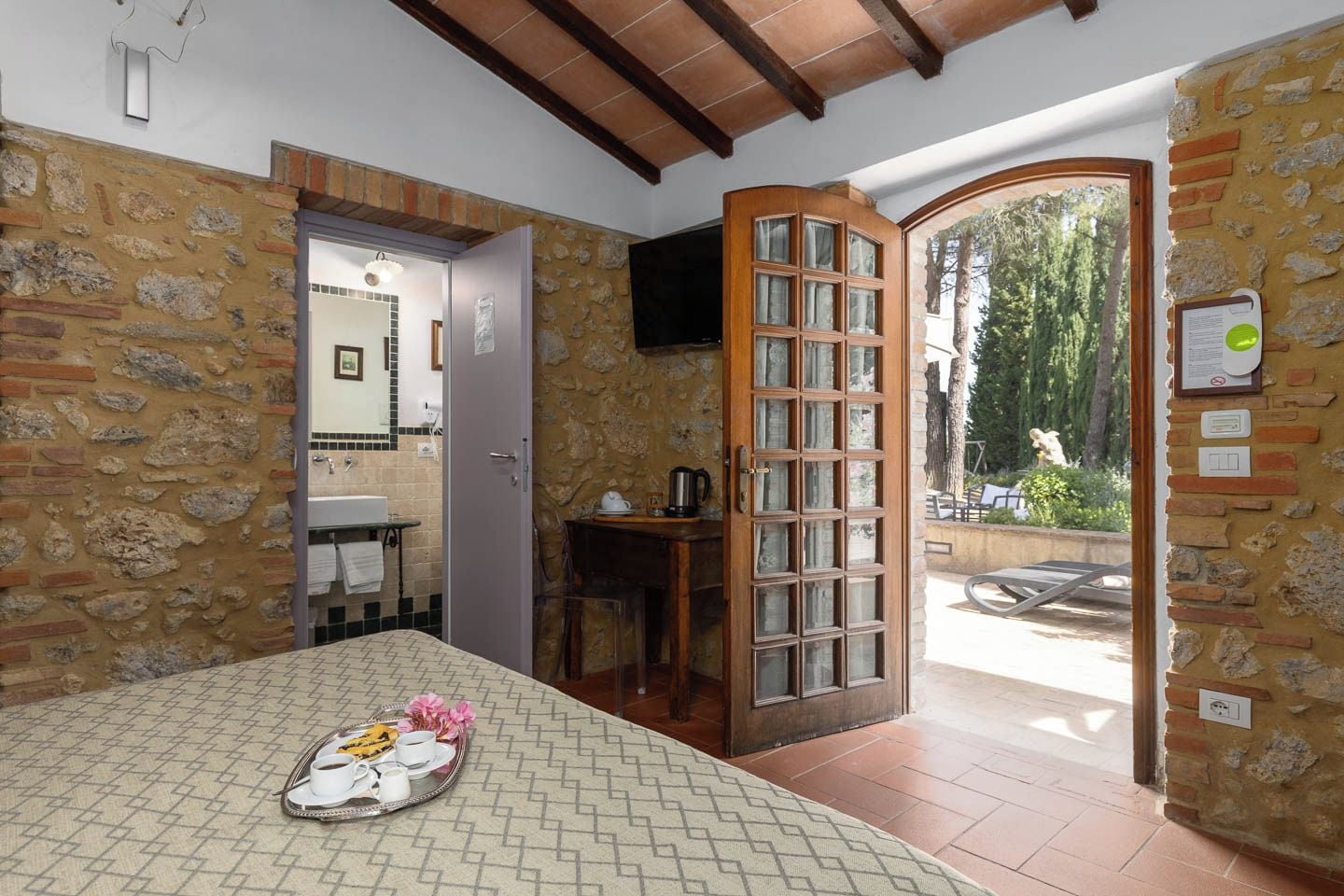 Una stanza con parete in pietra, porta in legno, pavimento piastrellato, un letto con un vassoio per la colazione e vista sull'esterno.