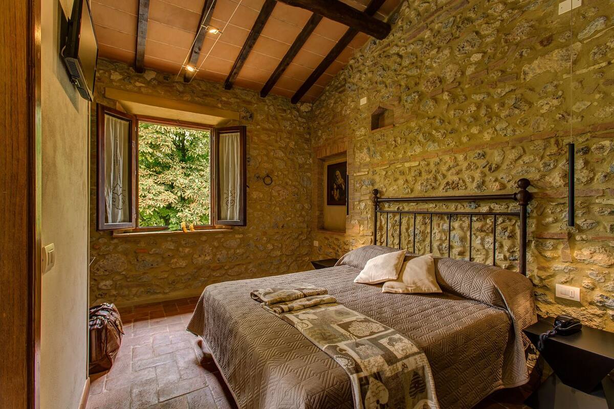 A bedroom with a stone wall, a bed with patterned bedding, a window with curtains, and a small table with a lamp.