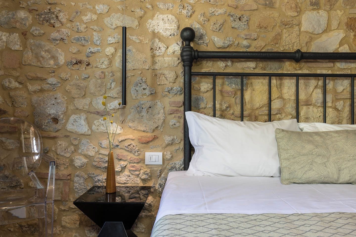 A bedroom with a stone wall, a metal headboard, a bed with white bedding, a bedside table with a vase, and a clear glass.