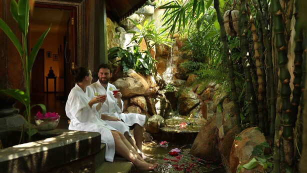 Dos personas sentadas junto a una fuente con plantas y rocas.