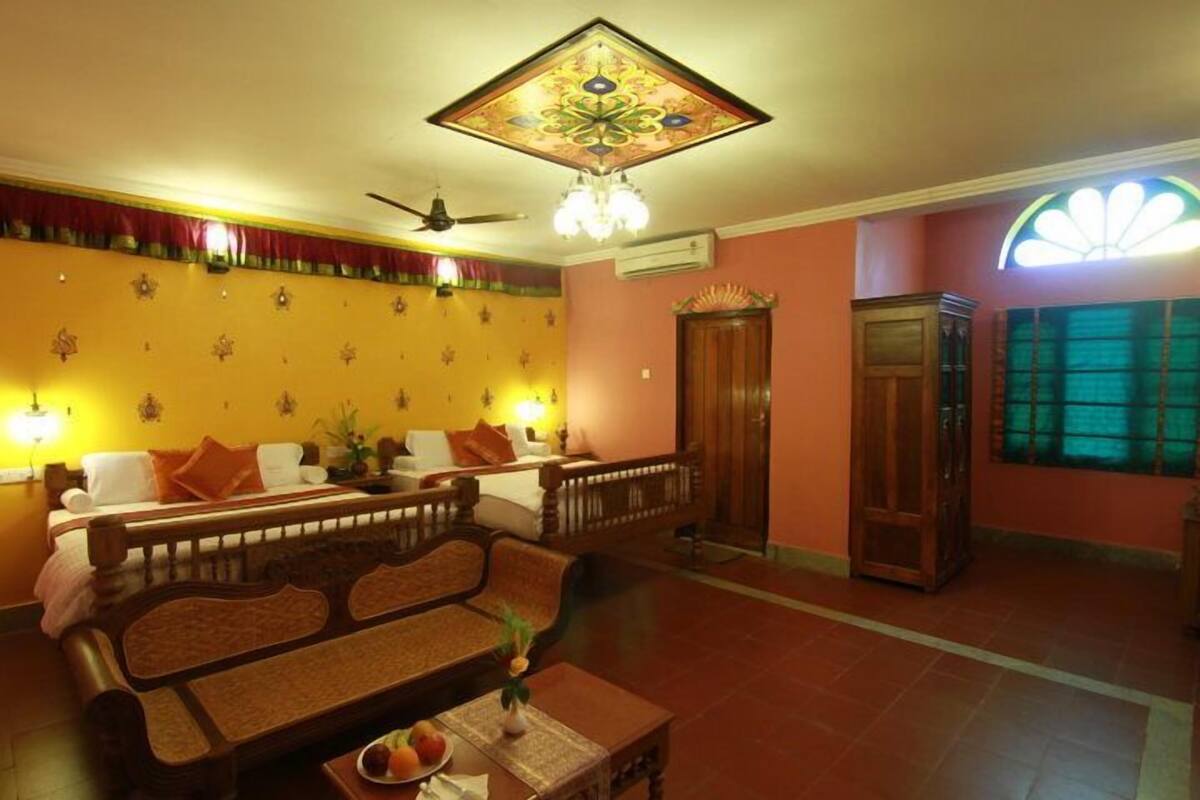 A hotel room with a sofa set, a dining table with a fruit bowl, a ceiling fan, and a decorative light fixture.