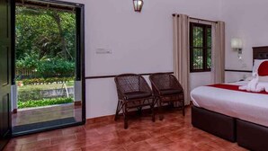 A hotel room with a bed, two chairs, a window with curtains, and a view of greenery outside. - Paradise Resorts (Kottayam)