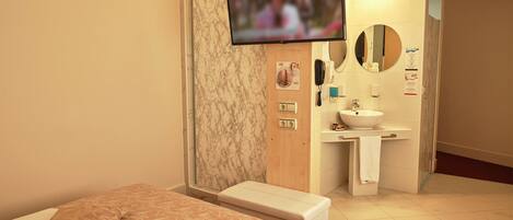 A hotel room with a bed, a bench, a TV mounted on the wall, a bathroom with a sink and mirror, and a wooden door.
