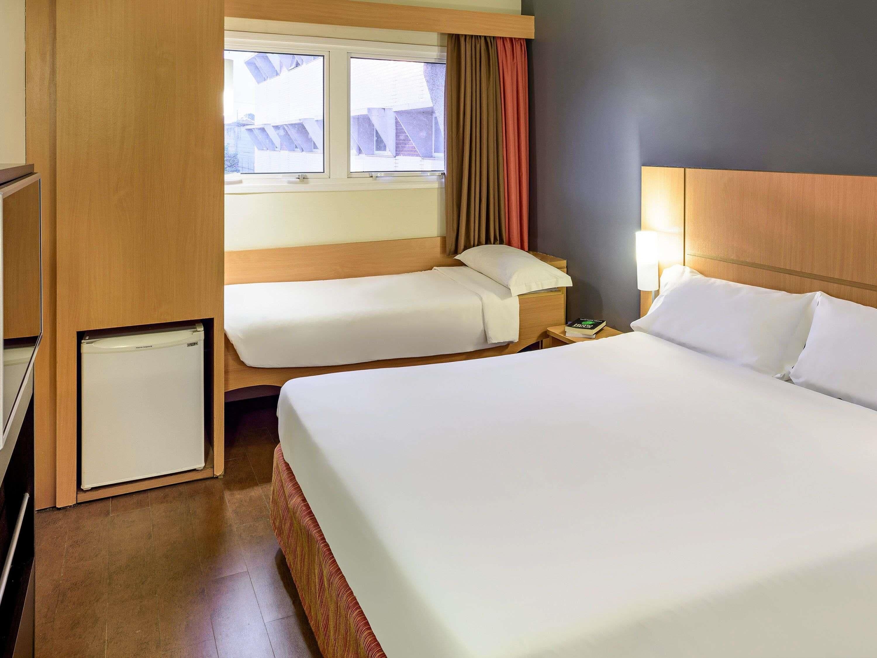 Une chambre d’hôtel avec deux lits, un petit réfrigérateur, une table de chevet avec une lampe et une fenêtre avec des rideaux.