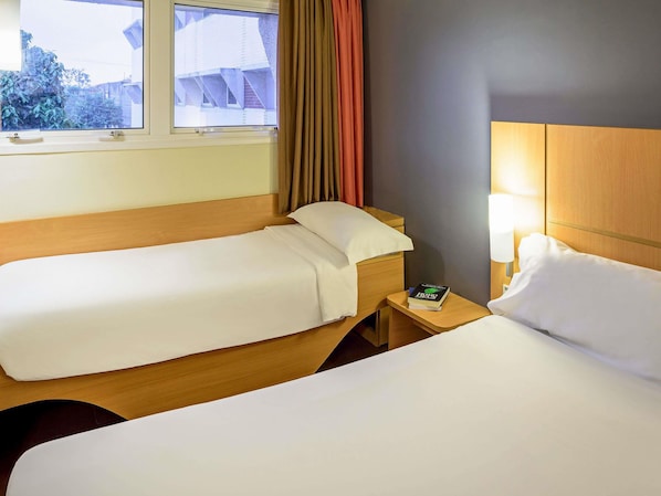 Una habitación de hotel con dos camas individuales, una mesita de noche con lámpara y una ventana con cortinas.