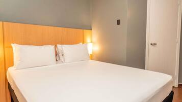 A hotel room with a bed, a wooden headboard, a wall-mounted light, and a door.