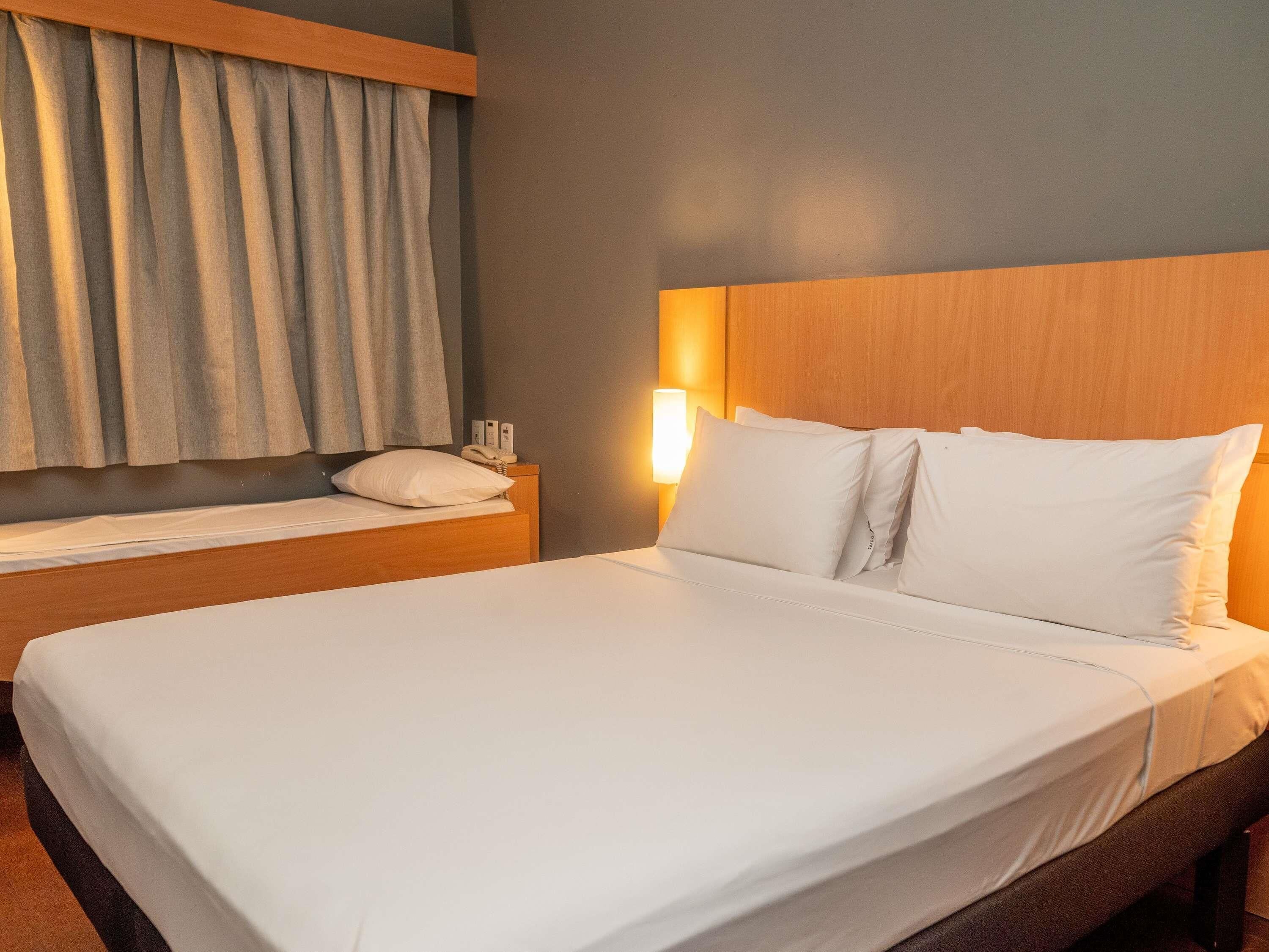 A hotel room with a bed, a nightstand, a lamp, and a window with curtains.