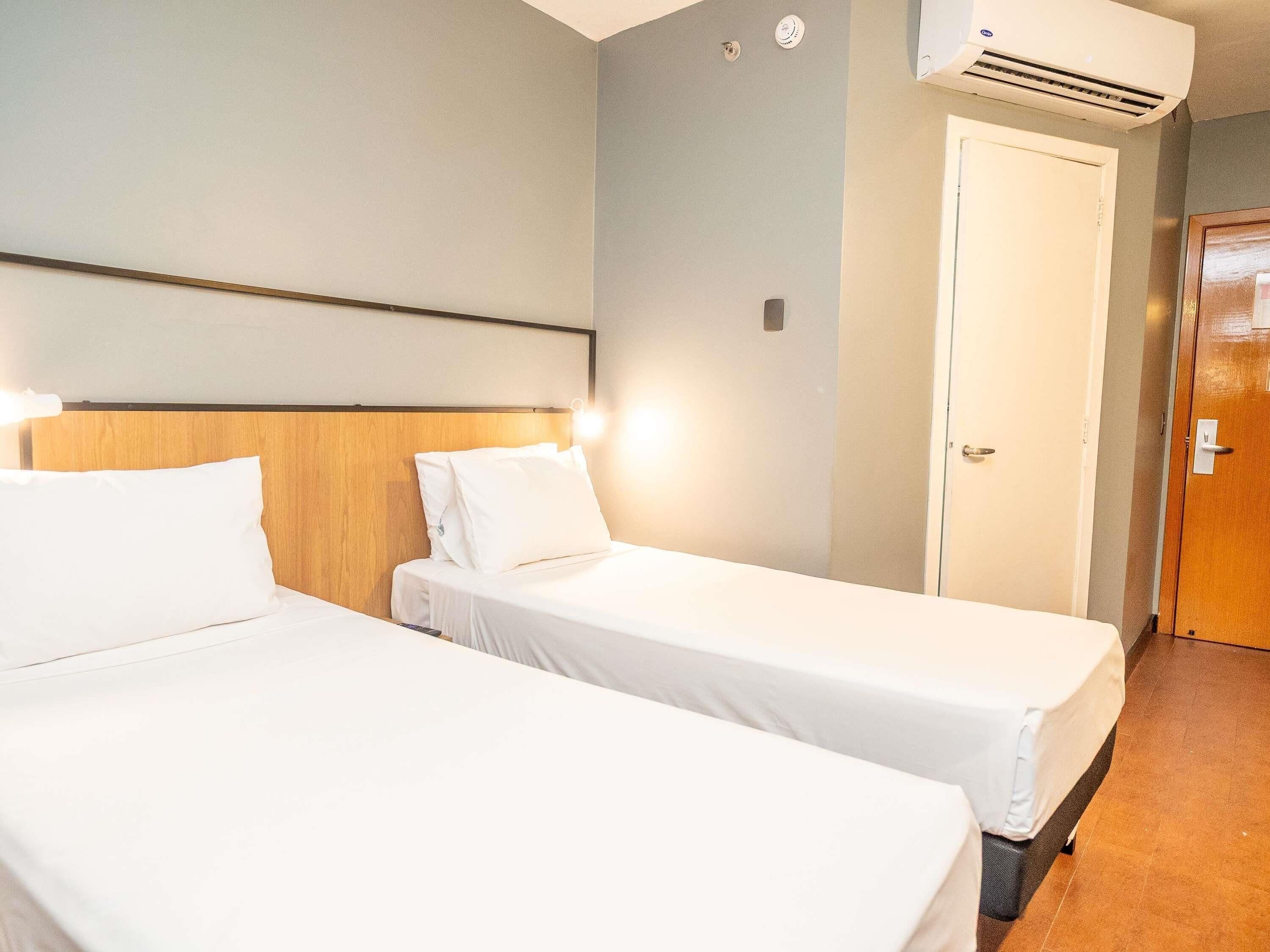 Habitación de hotel con dos camas individuales, cabecero de madera, unidad de aire acondicionado, puerta blanca y armario de madera.