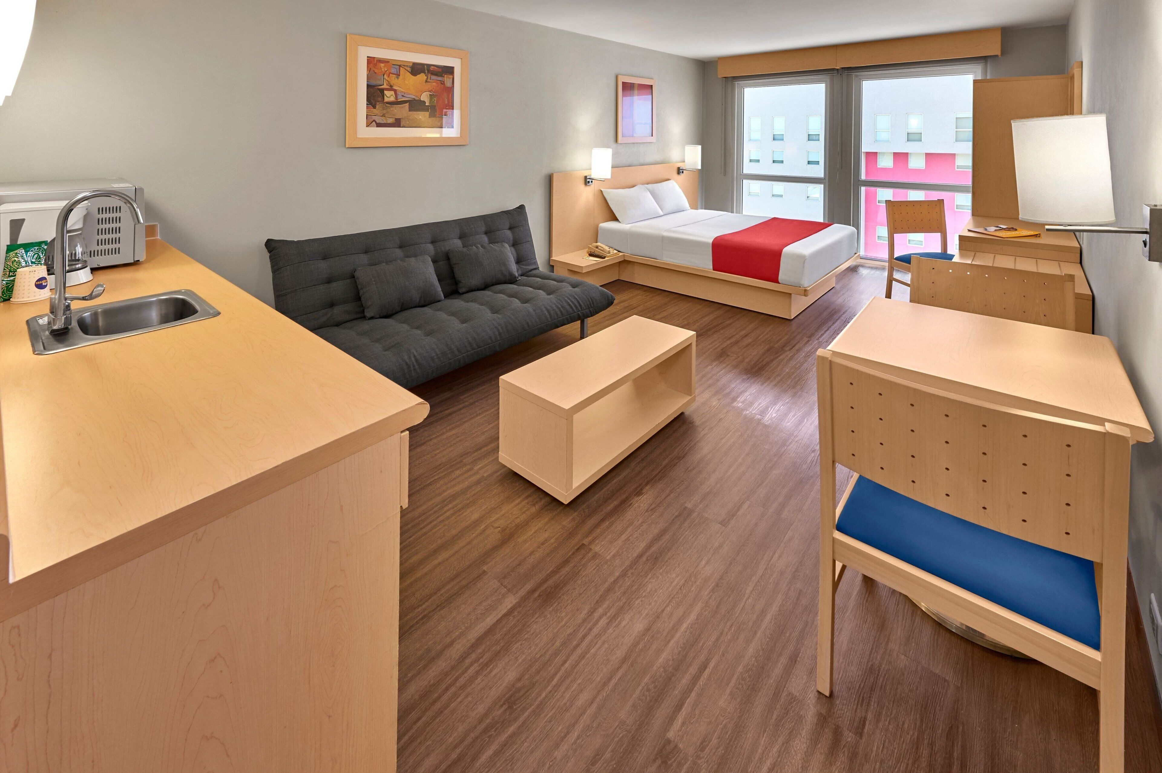 A hotel room with a kitchenette, a sofa, a bed with a red blanket, a desk with a chair, and a window with a view of buildings.