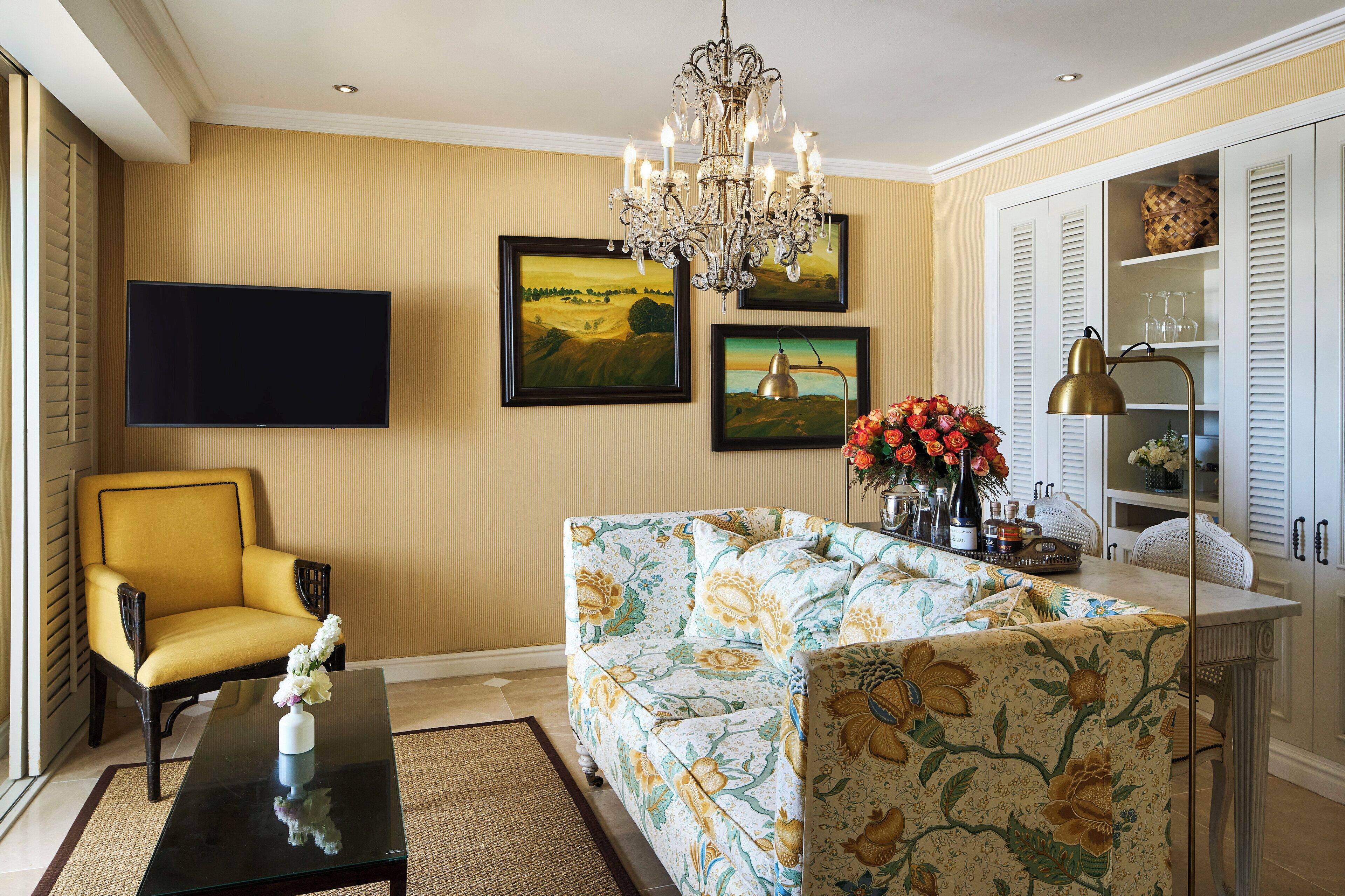 Una sala de estar con un sillón amarillo, un sofá con tapicería floral, un televisor de pantalla plana y una araña.