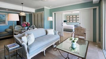 A well-lit room with a sofa, a glass coffee table, and a dining table. There is a door leading to another room with a bed and a wall decorated with framed pictures.
