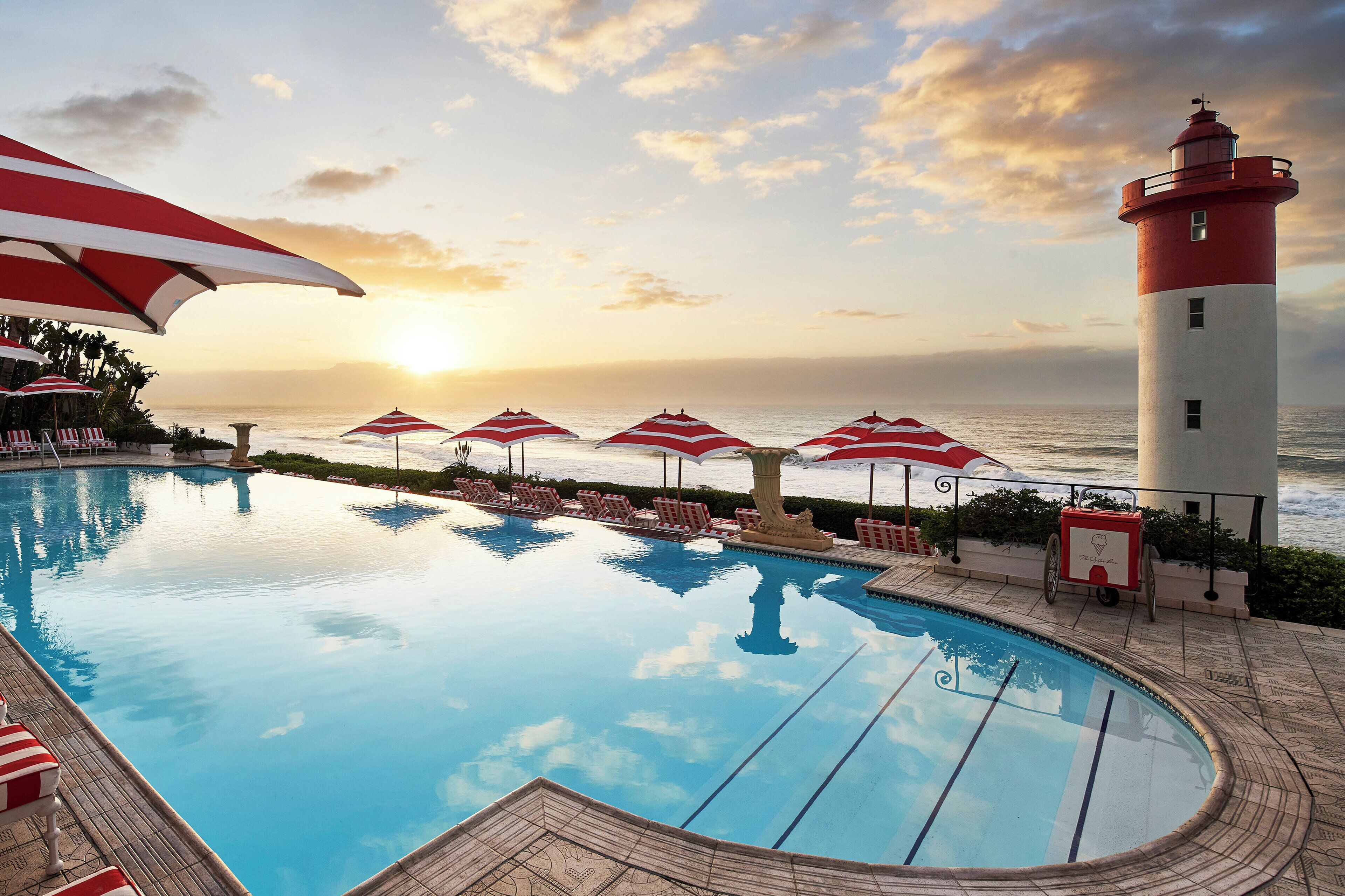 A swimming pool with lounge chairs and umbrellas, a lighthouse, and a sunset view.