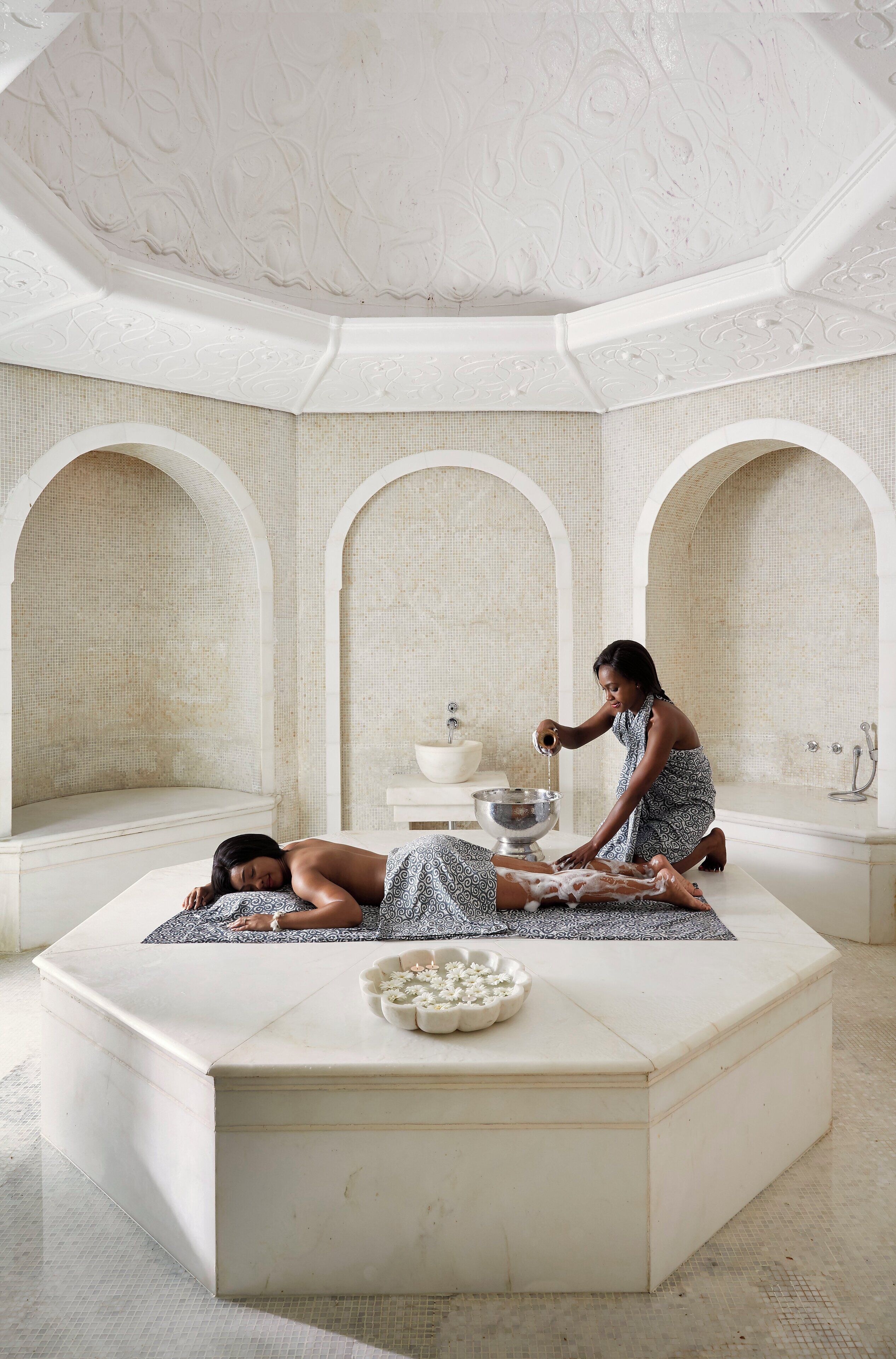 A spa room with a massage table, a person attending to another person, and a decorative bowl on a pedestal.