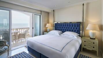 A spacious bedroom with a large bed, two bedside tables with lamps, a view of the sea through a sliding door, and a balcony with wicker furniture.