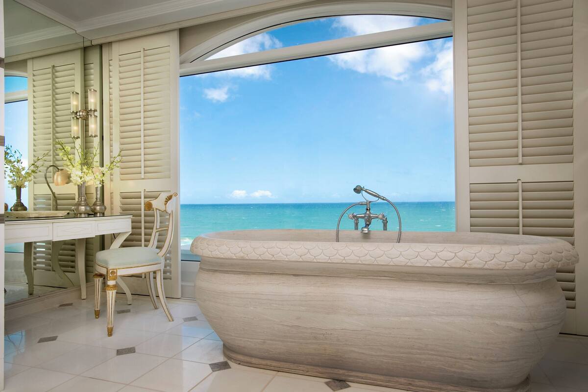 A bathroom with a large bathtub, a vanity with a mirror, and a chair. There is a view of the ocean through large windows.