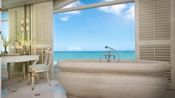 Un baño con una bañera grande, un tocador con espejo y una silla. Se ve el océano a través de ventanas amplias.