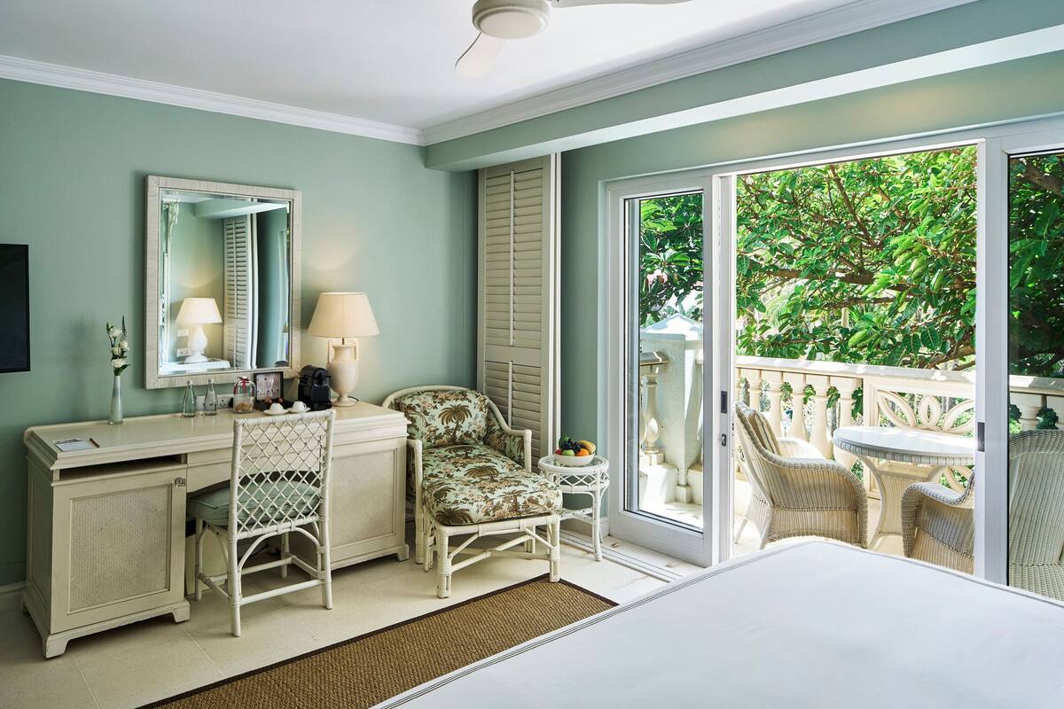 A bedroom with a desk, chair, bed, and a balcony with wicker furniture.