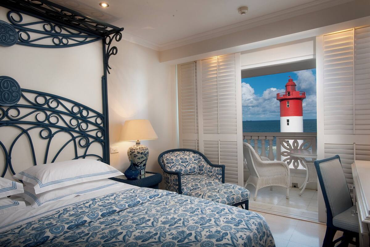 A bedroom with a bed, a nightstand, a lamp, and a view of a red lighthouse through a balcony.