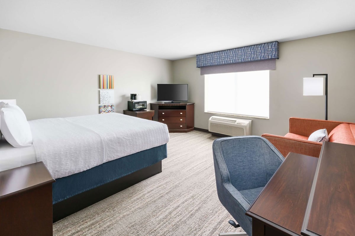 A hotel room with a bed, a desk, a chair, a TV, a dresser, and a window with blinds.
