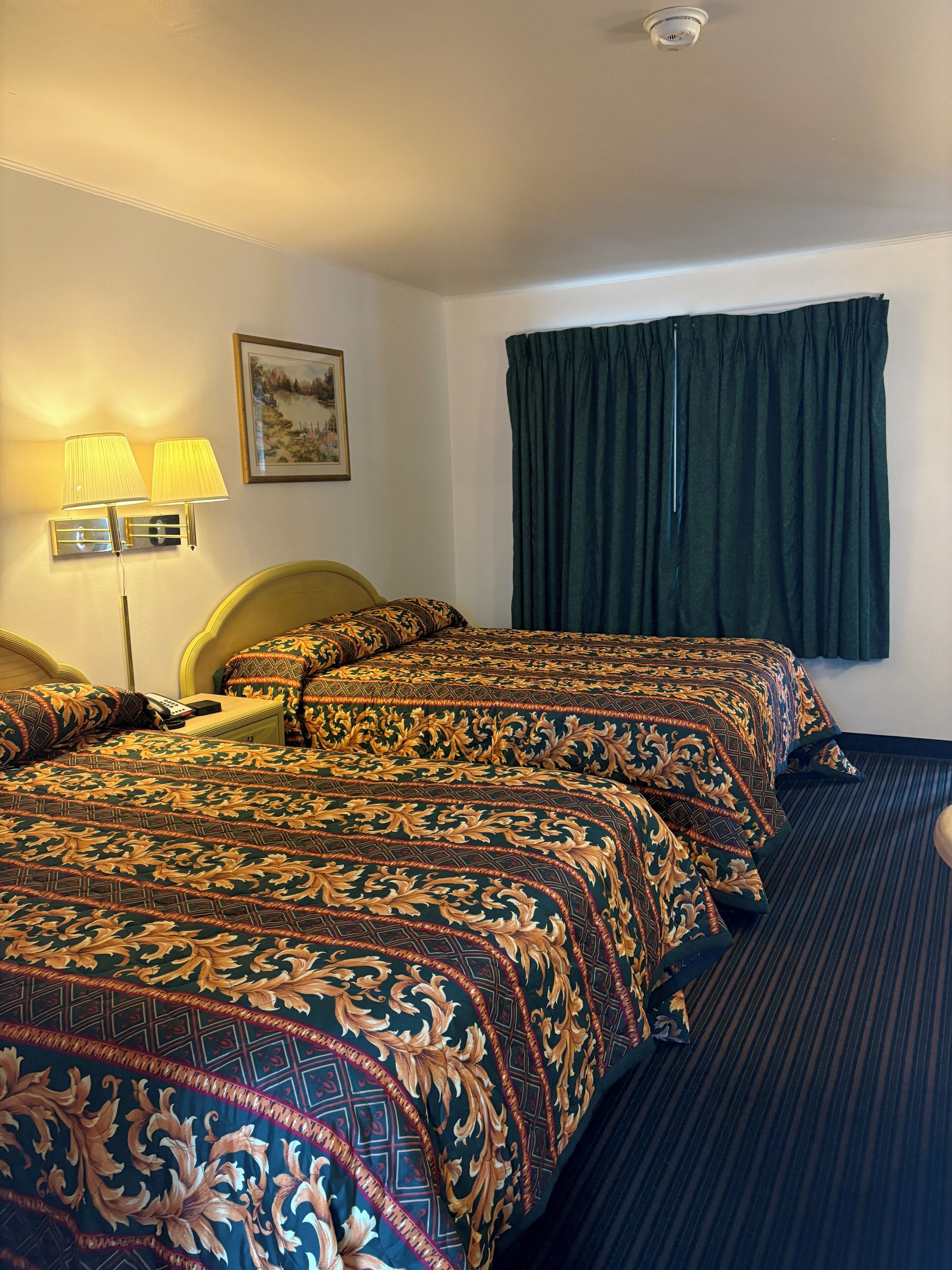 Una habitación de hotel con dos camas, una mesita de noche, una lámpara, un cuadro y una ventana con cortinas.