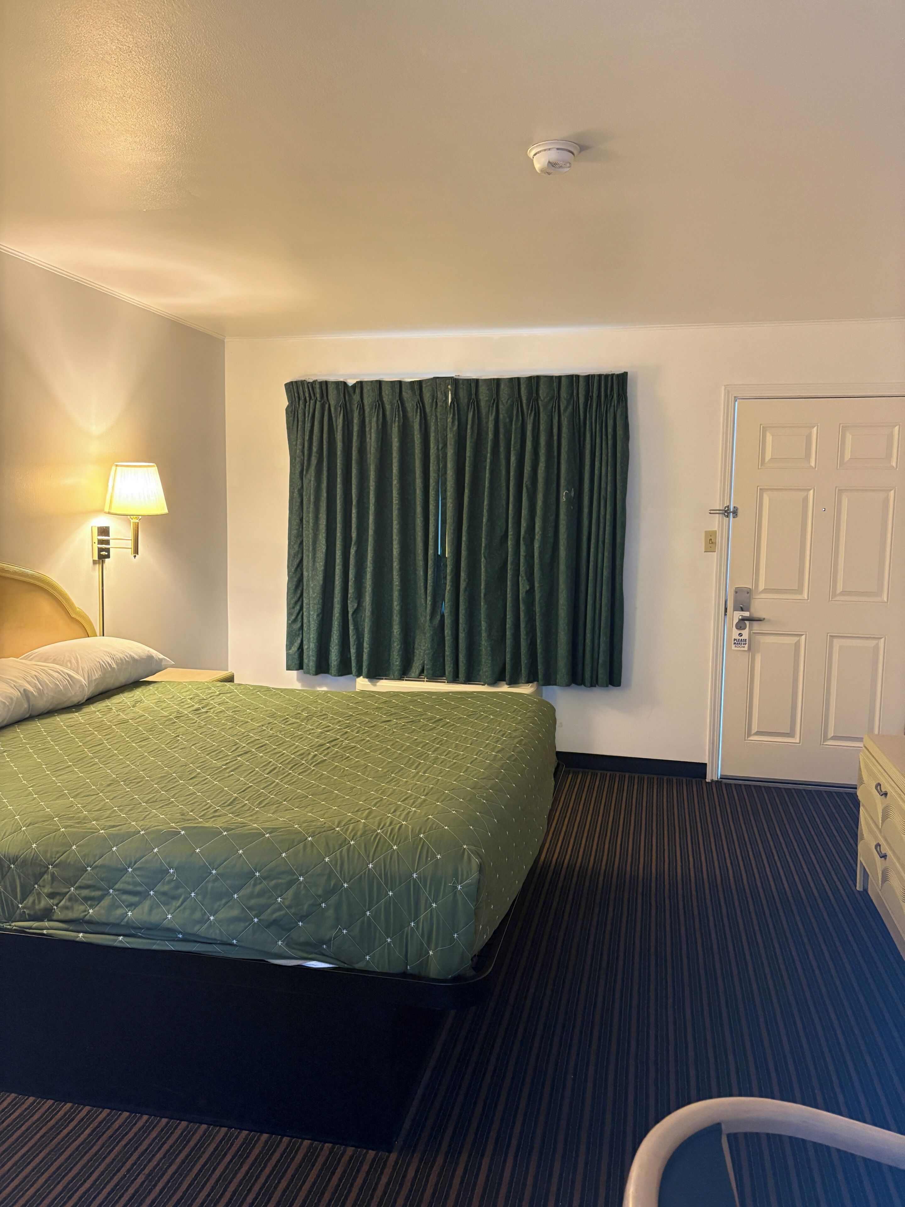 A hotel room with a bed, a nightstand, a lamp, a door, and a window with curtains.