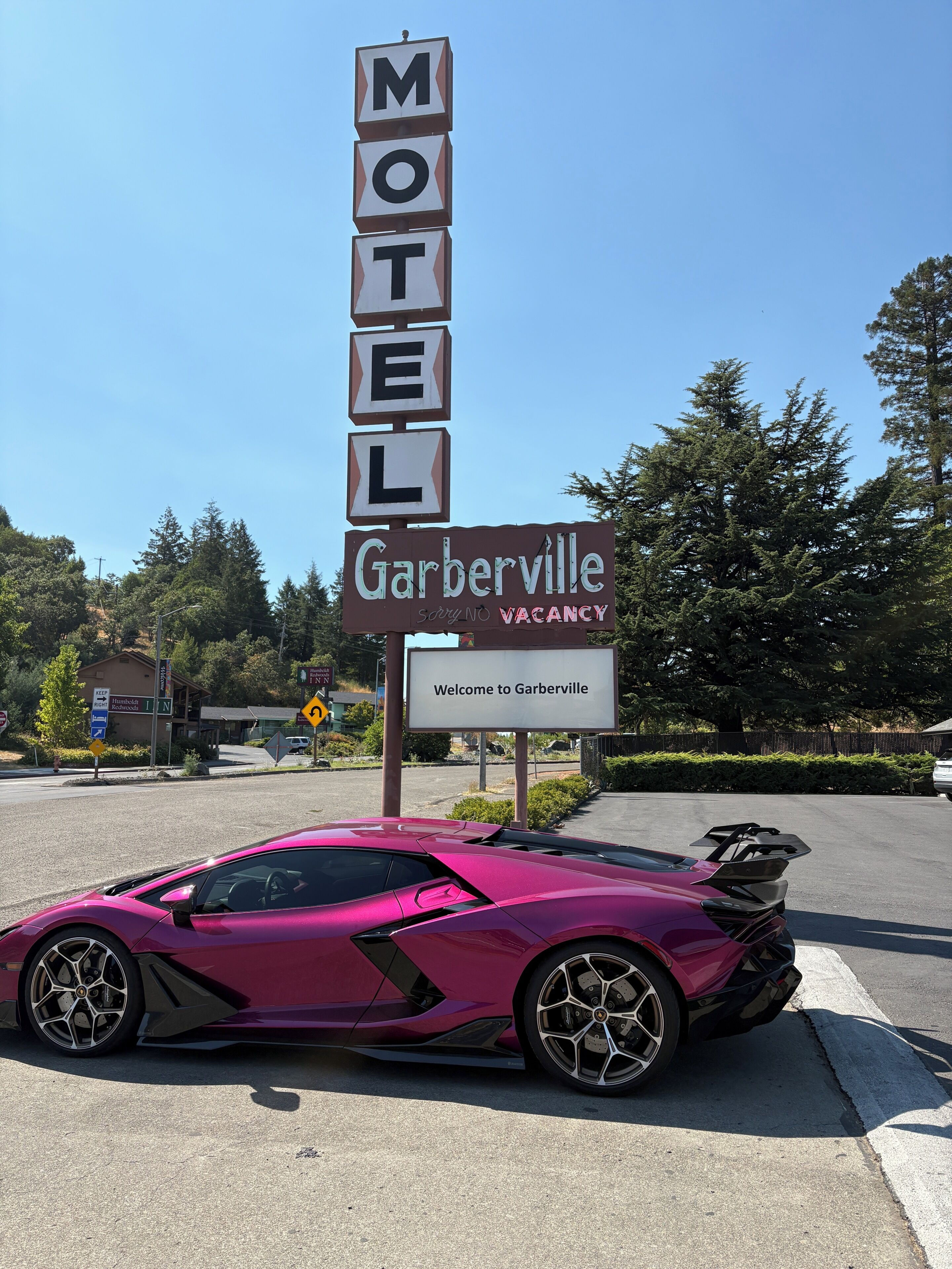 A motel sign with 'MOTEL' spelled out in blocks, a 'Garberville' sign below, and a pink sports car parked in front.