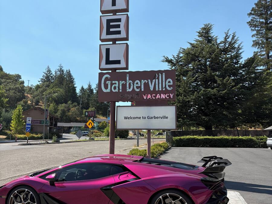 A motel sign with 'MOTEL' spelled out in blocks, a 'Garberville' sign below, and a pink sports car parked in front.