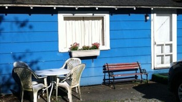 Un petit bâtiment bleu avec une porte et une fenêtre blanches, une table avec des chaises et un banc à l’extérieur.