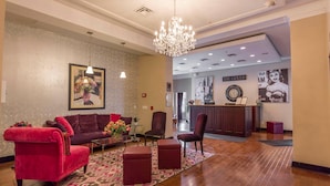Un hall d’hôtel avec un canapé en velours rouge, un pouf assorti et un tapis à motifs.